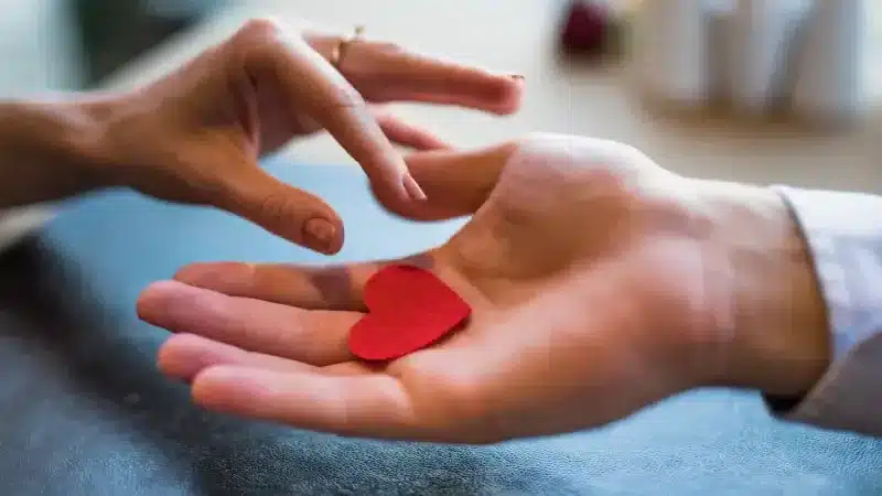 A hand extends towards another, which holds a small red felt heart, symbolizing love and connection between two people